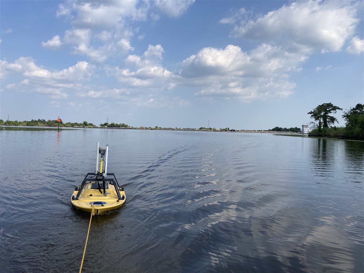 A small field vessel operating on open water near industrial shoreline conditions.